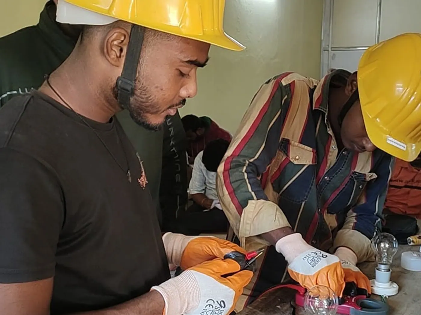 Two trainees in hard hats and safety gloves wiring a lamp circuit at an electrical bench